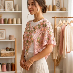May include: A pink crocheted shawl with floral embellishments. The shawl features a delicate openwork design and is adorned with three-dimensional flowers and green leaves. The garment is worn over a white top and paired with a beige skirt.