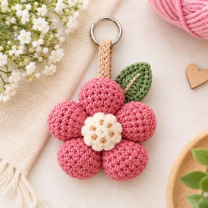 May include: A crocheted flower keyring featuring five rounded pink petals, a textured cream-coloured centre, and a single green leaf. The flower is attached to a beige crocheted loop and a silver-toned metal ring for secure attachment to items.