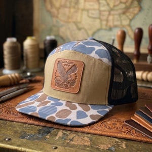 Puede incluir: Gorra de camionero con un panel frontal beige y una parte trasera de malla negra. La visera presenta un patrón de camuflaje azul, marrón y blanco. Un parche de cuero muestra un águila y el texto "1776-2026 AMERICA".