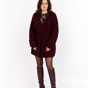 May include: A burgundy-colored, long-sleeved cardigan with a matching belt. The cardigan is paired with black tights and burgundy knee-high boots with pointed toes and high heels. A black lace top is visible underneath the cardigan.