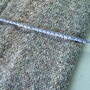 May include: Close-up of a light gray knitted fabric with a measuring tape laid across it. The tape shows measurements in inches. The fabric has a textured appearance with flecks of darker and lighter shades.