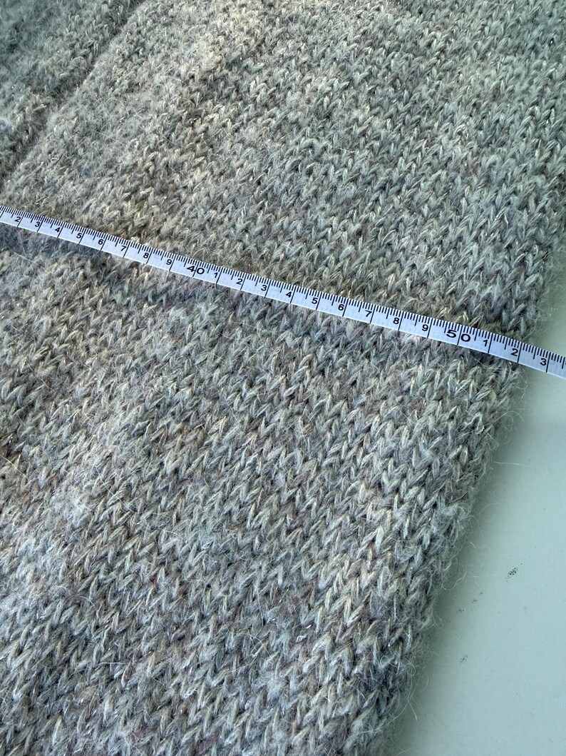 May include: Close-up of a light gray knitted fabric with a measuring tape laid across it. The tape shows measurements in inches. The fabric has a textured appearance with flecks of darker and lighter shades.