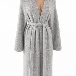 May include: A long, light gray knit cardigan with a V-neck and long sleeves. The cardigan features a matching belt that cinches at the waist. The soft, textured fabric drapes to the mid-calf.