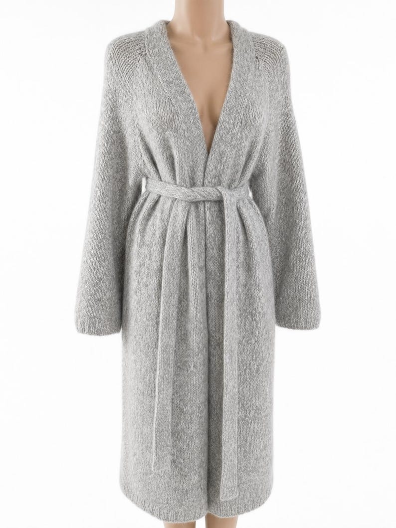 May include: A long, light gray knit cardigan with a V-neck and long sleeves. The cardigan features a matching belt that cinches at the waist. The soft, textured fabric drapes to the mid-calf.