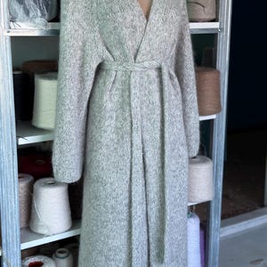 May include: A long, light gray knitted cardigan with a V-neck and a matching belt. The cardigan is displayed on a mannequin and appears to be made of a soft, textured yarn. The sleeves are long, and the cardigan falls to the ankles.