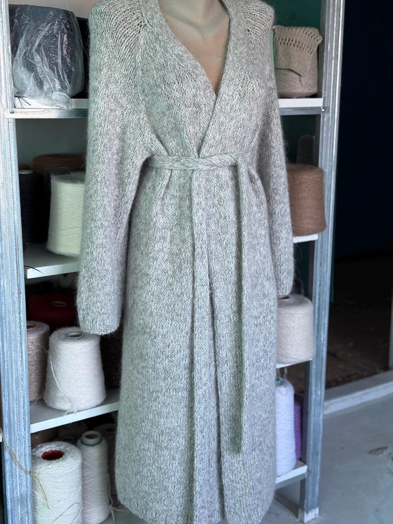 May include: A long, light gray knitted cardigan with a V-neck and a matching belt. The cardigan is displayed on a mannequin and appears to be made of a soft, textured yarn. The sleeves are long, and the cardigan falls to the ankles.
