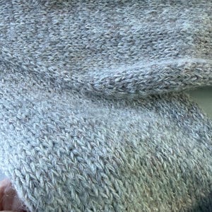 May include: Close-up of a light gray knitted fabric with a textured surface. The knitwear has a subtle shimmer, with strands of silver thread woven throughout. The fabric appears soft and cozy, suitable for a sweater or scarf.