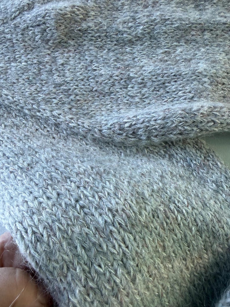 May include: Close-up of a light gray knitted fabric with a textured surface. The knitwear has a subtle shimmer, with strands of silver thread woven throughout. The fabric appears soft and cozy, suitable for a sweater or scarf.