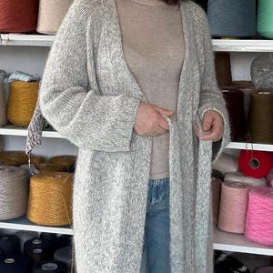 May include: A woman wearing a long, light gray knit cardigan over a beige top and blue jeans. Shelves behind her are filled with colorful yarn spools, suggesting a craft or textile theme.