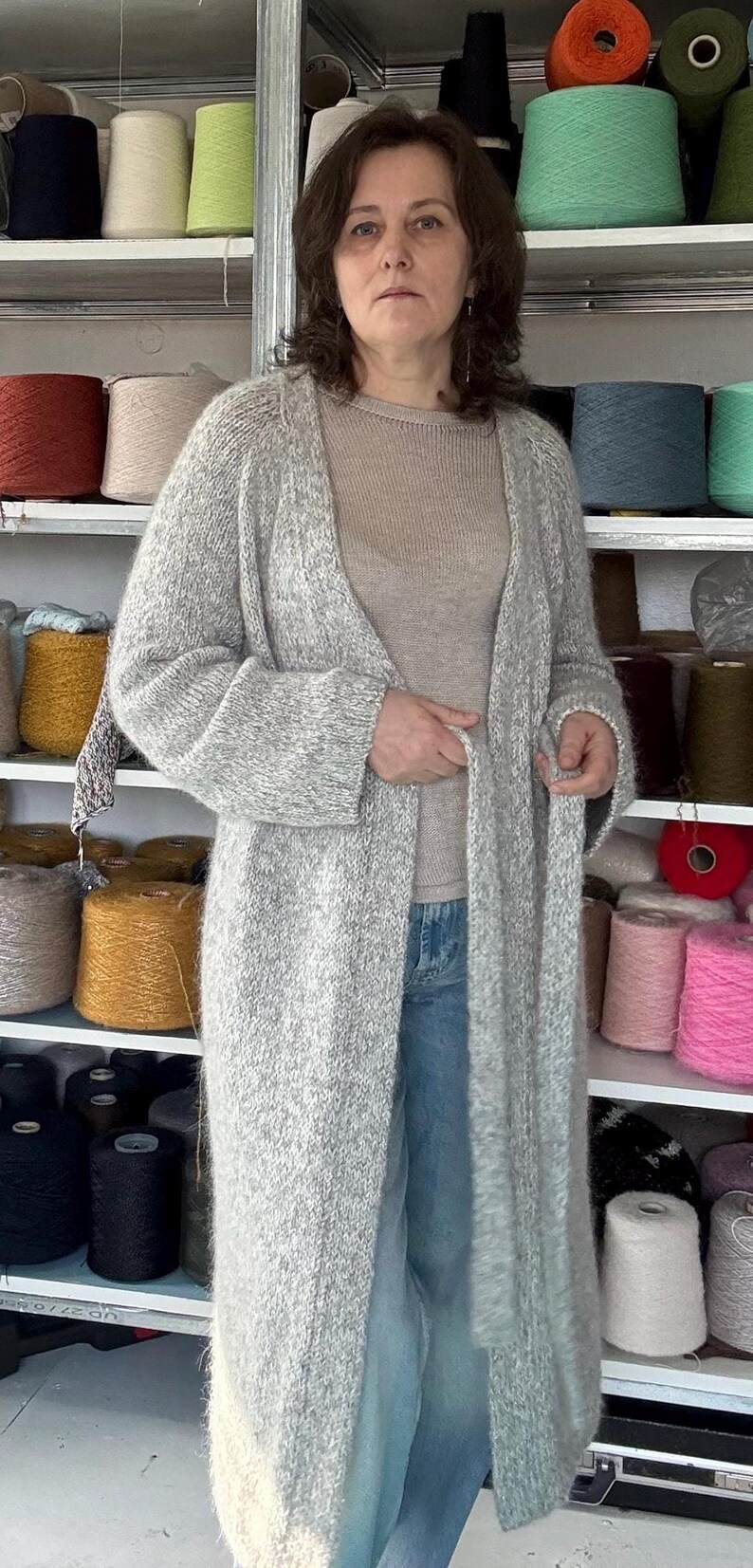 May include: A woman wearing a long, light gray knit cardigan over a beige top and blue jeans. Shelves behind her are filled with colorful yarn spools, suggesting a craft or textile theme.
