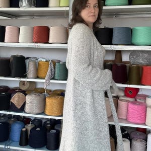 May include: A woman wearing a long, light gray cardigan with a matching belt stands in front of shelves filled with spools of yarn in various colors. The yarn colors include black, white, pink, and teal.