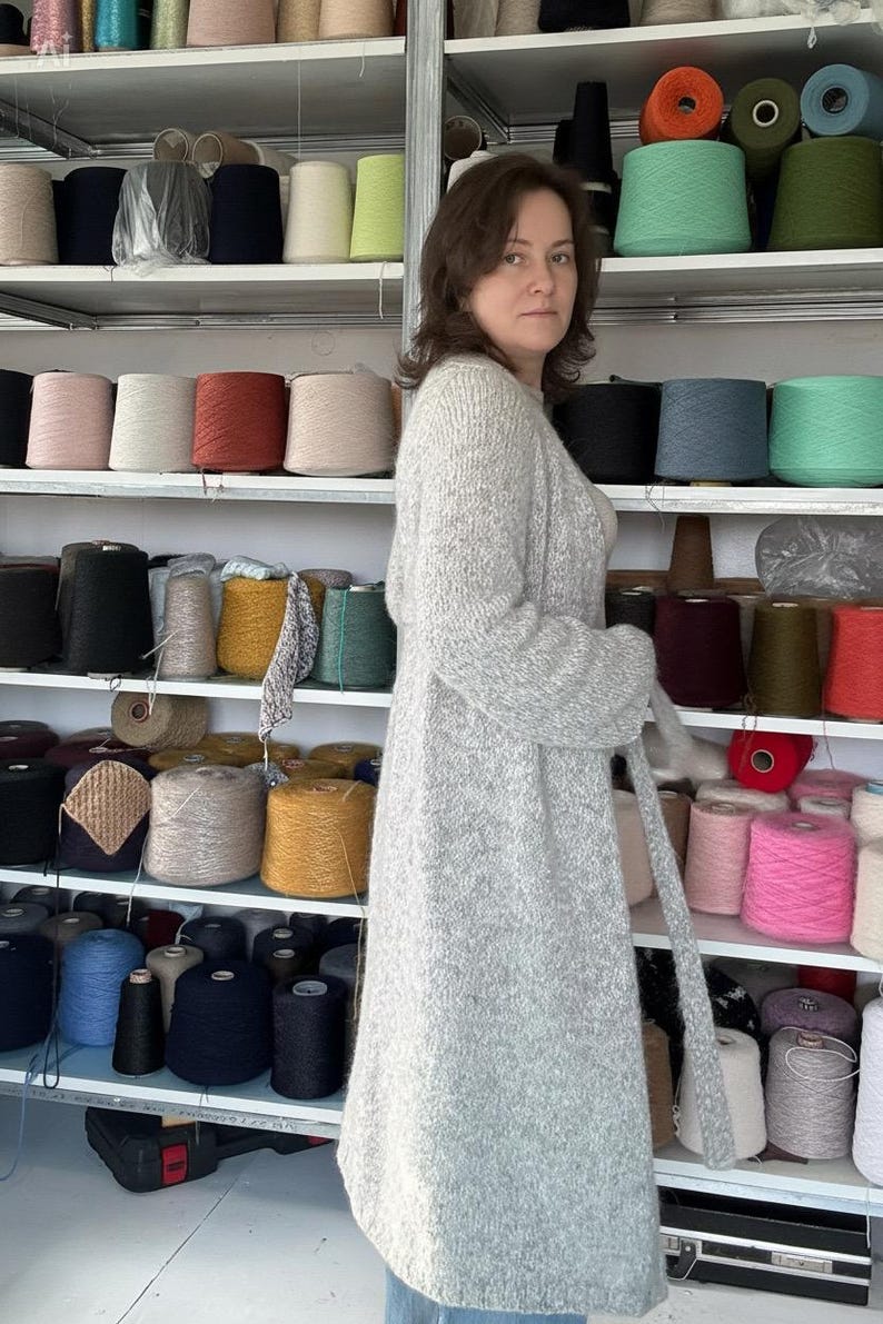 May include: A woman wearing a long, light gray cardigan with a matching belt stands in front of shelves filled with spools of yarn in various colors. The yarn colors include black, white, pink, and teal.