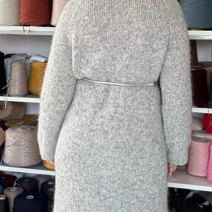 May include: A light gray, long knitted coat with a silver belt cinching the waist. The coat is made of a thick, textured yarn. Shelves in the background display various spools of yarn in a range of colors, suggesting a craft or textile theme.