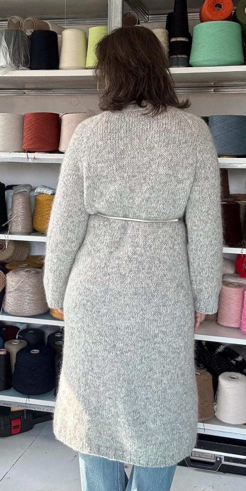 May include: A light gray, long knitted coat with a silver belt cinching the waist. The coat is made of a thick, textured yarn. Shelves in the background display various spools of yarn in a range of colors, suggesting a craft or textile theme.