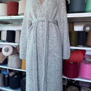 May include: A long, light gray knit cardigan with a V-neck and a matching belt. The cardigan is displayed on a mannequin. Shelves in the background are filled with spools of yarn in various colors, including red, pink, and blue.