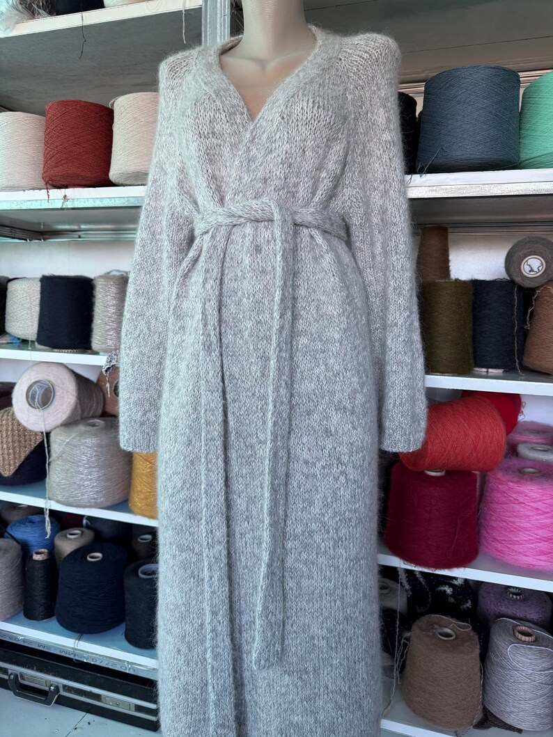 May include: A long, light gray knit cardigan with a V-neck and a matching belt. The cardigan is displayed on a mannequin. Shelves in the background are filled with spools of yarn in various colors, including red, pink, and blue.