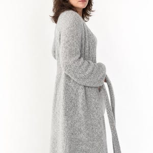 May include: A long, light gray knit cardigan with a matching belt. The cardigan is knee-length and has long sleeves. The model is wearing blue jeans. The cardigan is made of a soft, textured material.