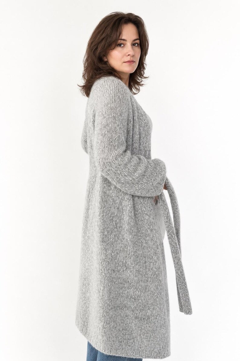May include: A long, light gray knit cardigan with a matching belt. The cardigan is knee-length and has long sleeves. The model is wearing blue jeans. The cardigan is made of a soft, textured material.