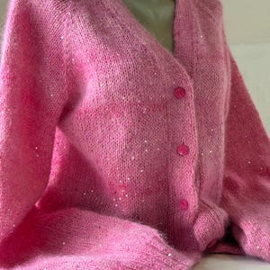 May include: A bright pink V-neck cardigan sweater featuring a fuzzy, textured knit fabric embellished with small, scattered silver sequins. The garment includes long sleeves, ribbed cuffs, and a front button closure with three matching pink buttons.