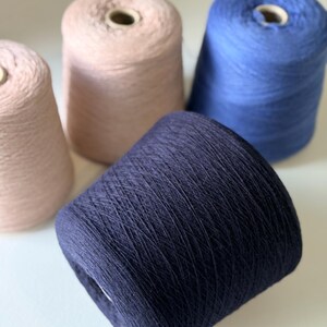 May include: Close-up of four spools of yarn in shades of blue and pink. The yarn is tightly wound on cardboard cores. The colors include a deep navy, a light blue, and two shades of pink. The yarn appears soft and suitable for knitting or crocheting projects.