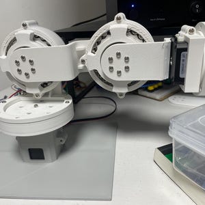 以下が含まれることがあります： 複数の関節と円形コンポーネントを備えた白いロボットアーム。ネジで固定されています。アームは灰色のベースに取り付けられており、プラスチックまたは同様の素材で作られているようです。設計は機械的または工学的な用途を示唆しています。