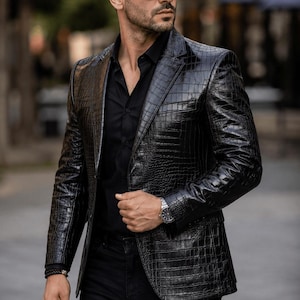 Op de afbeelding: Een zwarte leren blazer met een krokodillenleertextuur. De blazer wordt gedragen over een zwart overhemd en een zwarte broek. De persoon draagt een zwarte zonnebril en een zilveren horloge.