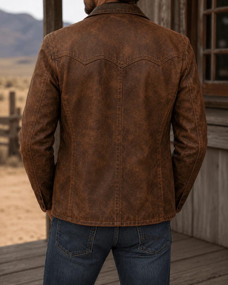 May include: A brown leather jacket with a distressed finish. The jacket features a classic collar, buttoned cuffs, and a Western-style yoke on the back. The person is wearing blue jeans. The background shows a wooden structure and a landscape.