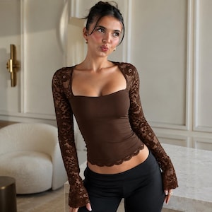 May include: A long-sleeved brown top featuring a square neckline and intricate lace sleeves. The garment has a fitted bodice with a scalloped lace hemline that extends along the bottom edge, providing a textured contrast to the solid fabric.
