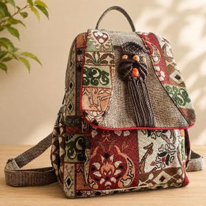May include: A backpack featuring a multi-colored woven tapestry fabric with geometric and floral patterns in shades of red, green, and tan. The bag includes a top flap with a dark brown macrame ornament, a top handle, and adjustable shoulder straps.