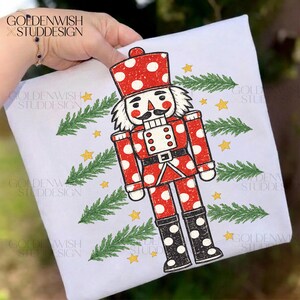 May include: A white t-shirt featuring a graphic of a nutcracker with a red and white polka dot uniform, black boots with white dots, and a black moustache. The design is surrounded by green pine branch illustrations and small yellow stars.