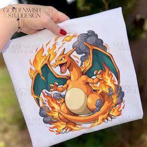 May include: A white t-shirt featuring a graphic illustration of a dragon-like creature with orange skin, teal wings, and a cream-colored belly. The creature is surrounded by bright orange and yellow flames and dark grey smoke clouds.
