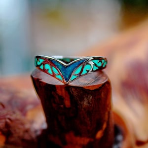 May include: A silver ring with a turquoise and blue inlay. The ring has a V-shaped design with intricate scrollwork and is displayed on a piece of wood. The ring's design is highlighted by the contrast of the silver and the vibrant colors.