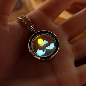 May include: A silver locket necklace with a clear glass insert. Inside the glass is a yellow sun and blue clouds.