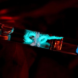 May include: A clear glass tube with a silver cap and a blue glowing substance inside. The tube is lying on a dark red surface.