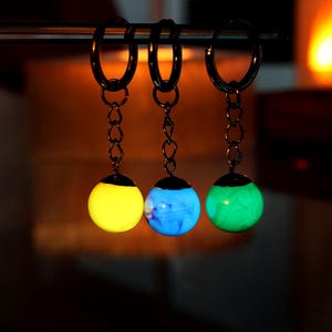 Könnte beinhalten: Drei Schlüsselanhänger, die im Dunkeln leuchten, mit unterschiedlich farbigen Kugeln. Die Kugeln sind gelb, blau und grün.