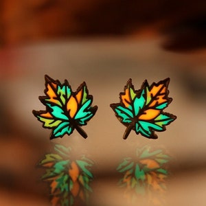 May include: A pair of small, colourful leaf-shaped earrings. The leaves are green, yellow and orange. The earrings are reflected in a shiny surface.