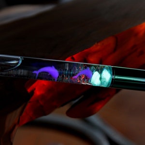 May include: A clear pen with a rainbow iridescent finish. The pen contains a purple dolphin and white glitter inside the clear barrel.