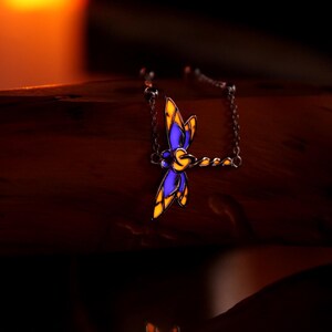 May include: A dragonfly pendant necklace with a silver chain. The dragonfly has blue and yellow stained glass-style wings. The necklace is displayed on a dark wooden surface, with a soft, warm light in the background.