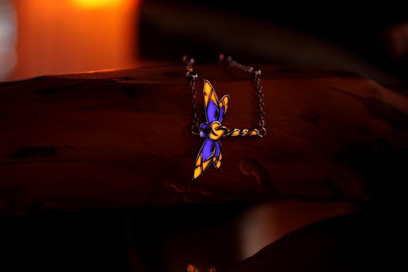 May include: A dragonfly pendant necklace with a silver chain. The dragonfly has blue and yellow stained glass-style wings. The necklace is displayed on a dark wooden surface, with a soft, warm light in the background.
