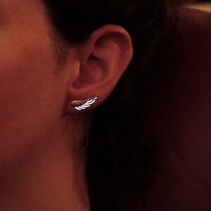 K&ouml;nnte beinhalten: Nahaufnahme eines Ohres mit einem kleinen silbernen Fl&uuml;gelohrring. Der Ohrring ist ein schlichtes Design mit einer zarten Fl&uuml;gelform.