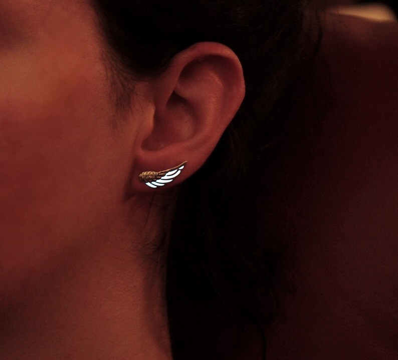 K&ouml;nnte beinhalten: Nahaufnahme eines Ohres mit einem kleinen silbernen Fl&uuml;gelohrring. Der Ohrring ist ein schlichtes Design mit einer zarten Fl&uuml;gelform.