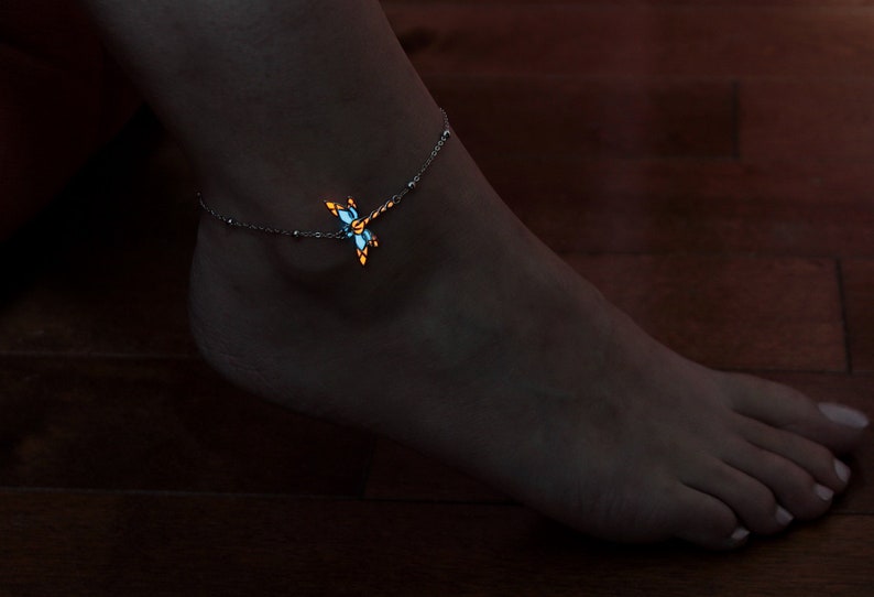 May include: Ankle bracelet featuring a dragonfly design. The dragonfly has blue and orange accents and appears to glow. The bracelet is silver-toned and delicate, with small beads. The image is set against a dark background.