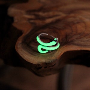 Puede incluir: Un anillo de serpiente de plata que brilla en verde. El anillo está sobre una superficie de madera.
