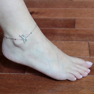 May include: A silver anklet with a dragonfly charm and small beaded accents. The anklet is worn on an ankle, with the foot resting on a wooden surface. The dragonfly charm has intricate details.