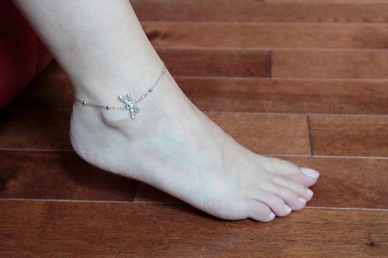 May include: A silver anklet with a dragonfly charm and small beaded accents. The anklet is worn on an ankle, with the foot resting on a wooden surface. The dragonfly charm has intricate details.