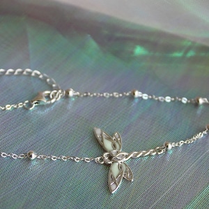 May include: A delicate silver anklet featuring a dragonfly charm. The anklet has a fine chain with small, round beads and an adjustable clasp. The dragonfly charm has white enamel detailing. The background is a shimmering, iridescent fabric.
