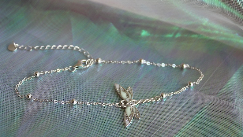 May include: A delicate silver anklet featuring a dragonfly charm. The anklet has a fine chain with small, round beads and an adjustable clasp. The dragonfly charm has white enamel detailing. The background is a shimmering, iridescent fabric.