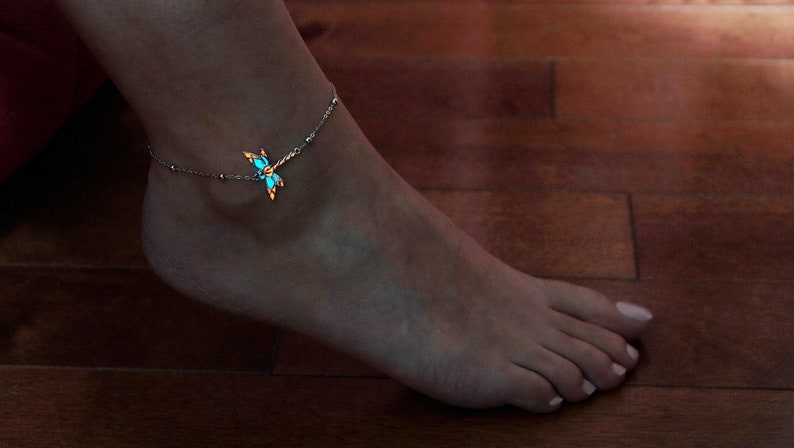 May include: A silver anklet with a dragonfly charm. The dragonfly has turquoise-colored accents that glow. The anklet is worn on a foot, with the background showing a wooden floor.