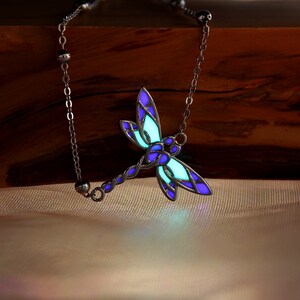 May include: A silver-toned dragonfly pendant necklace. The dragonfly features a stained-glass design with blue and purple wings, and a glowing green center. The necklace is displayed on a wooden surface.