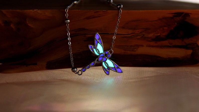 May include: A silver-toned dragonfly pendant necklace. The dragonfly features a stained-glass design with blue and purple wings, and a glowing green center. The necklace is displayed on a wooden surface.